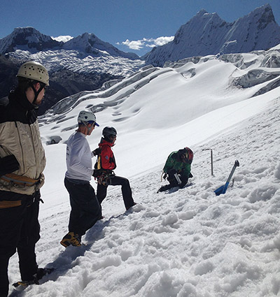 Four students dressed in winter gear on snow-covered mountain with big peaks in the background. One student is digging into the snow while the other three watch.