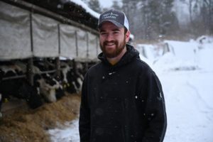 Man smiling, standing in lightly falling snow with cows in background.