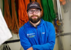 A man with glasses and a beard, smiling, looking at camera, with rock climbing equipment in background.