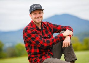 A young man wearing a hat and a red flannel, sitting outdoors, smiling, with mountains in background.