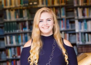 A young lady with long, blonde hair, smiling, looking at camera, with bookshelves in background.