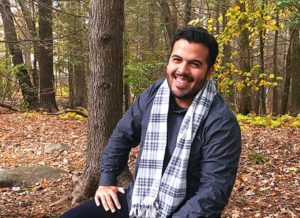 A man wearing a scarf with dark hair and a beard, smiling at the camera, trees in background.