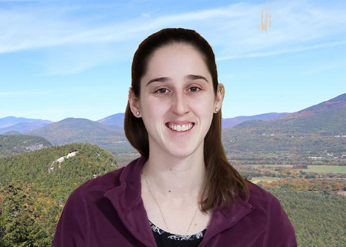 A young woman wearing a purple shirt, smiling, looking at camera, with mountains in background.