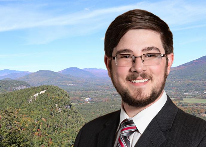 A young man with a beard and glasses, smiling, looking at camera, with mountains in background.