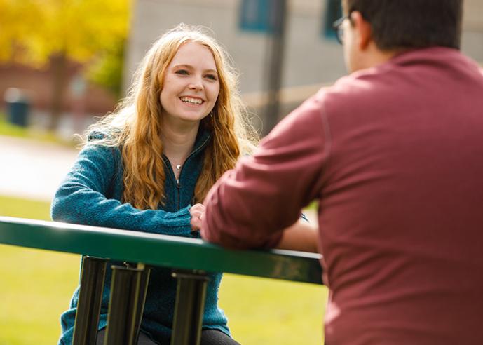 A girl sitting at an outdoor table, smiling, looking at the person sitting across from her.