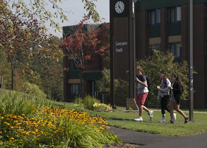 Students walking along a path, outside a college campus building, lined with trees.