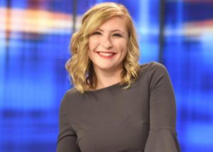 A young woman with blonde hair, smiling, looking at camera, with blue background.