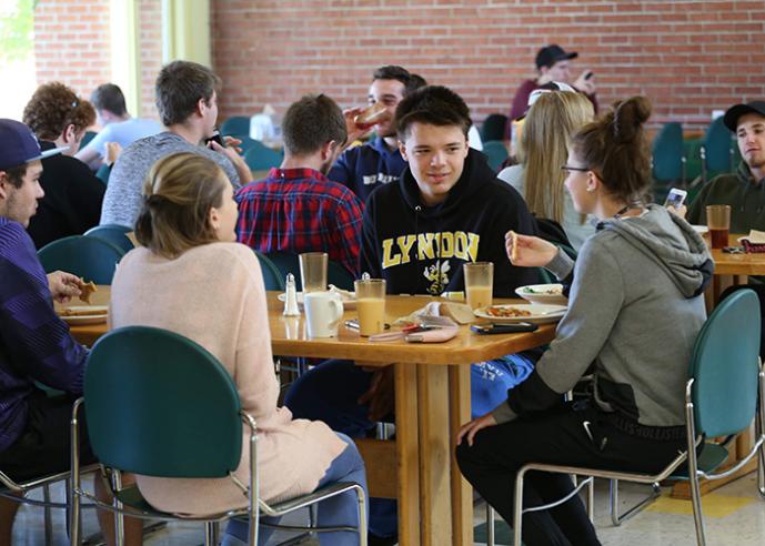 Students sit at a table in a cafeteria, eating lunch and talking.