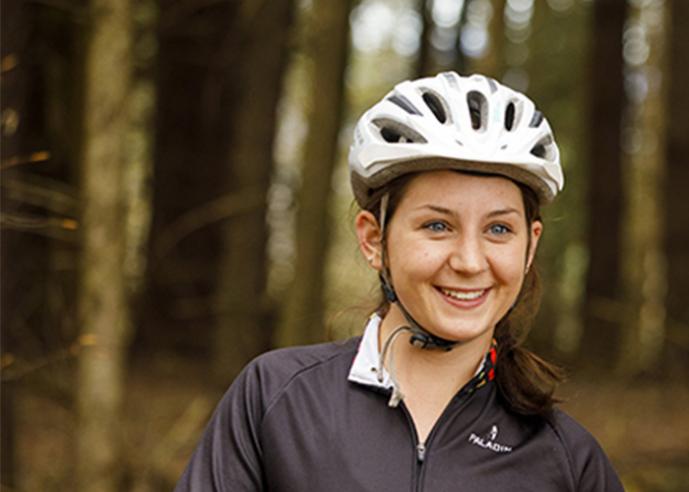 A young woman with a ponytail, wearing a bike helmet, smiling, with trees in background.