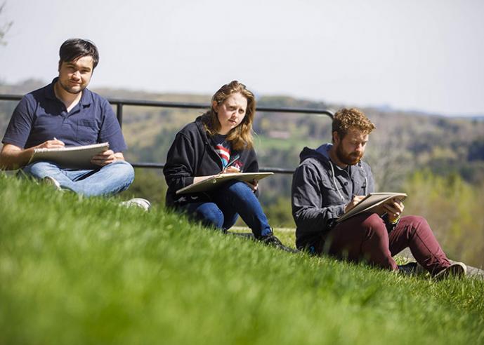 Three students sitting in grass, sketching in notebooks.
