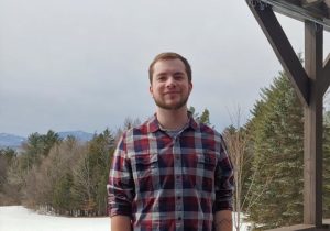 A young man wearing a flannel in cold weather, smiling, with trees in background.