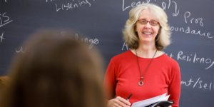 A female teacher wearing glasses and a red shirt, smiling to her class, with chalkboard in background.