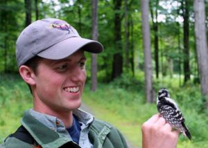 A man wearing a baseball cap, smiling at woodpecker that is perched on his hand.