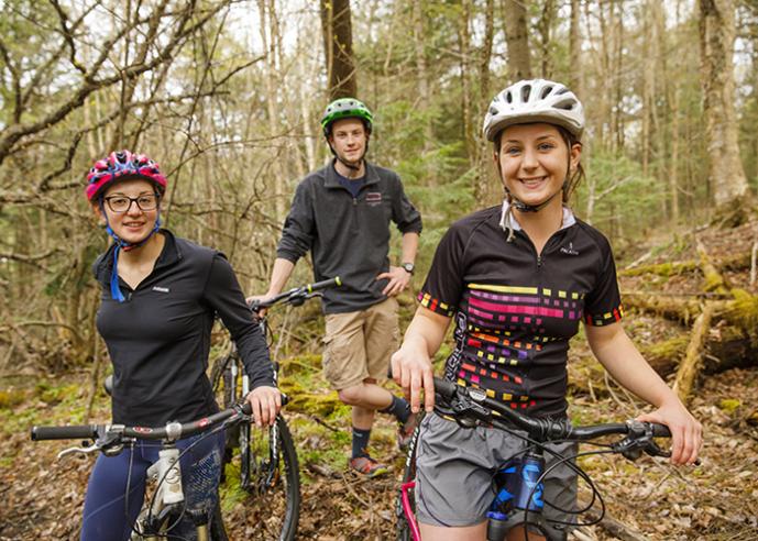 Three people with bikes wearing helmets, standing, smiling, looking at camera.