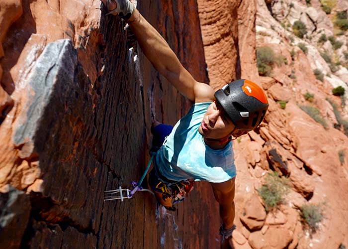 A young man in a helmet dangling by one hand from a desert cliff.