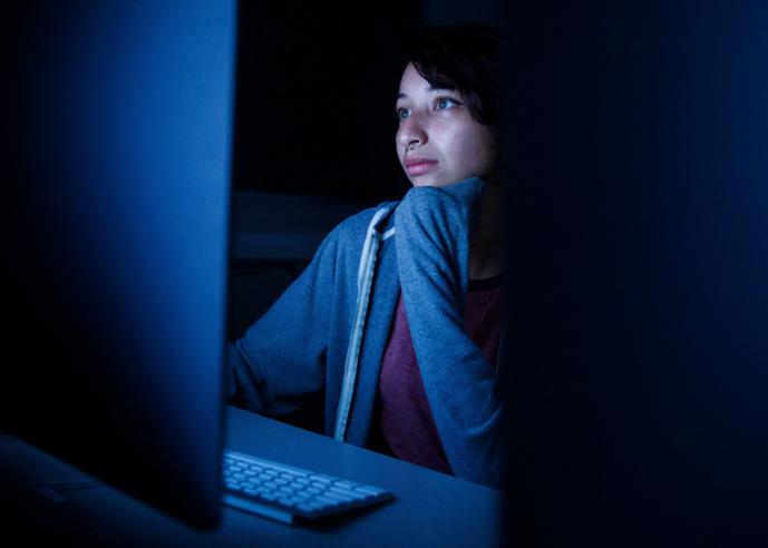 A student wearing a sweatshirt looks into a computer screen in a dark room.