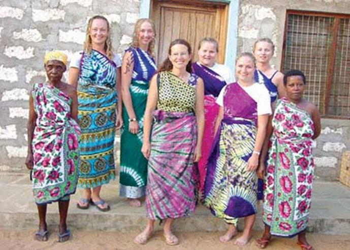 A group of women wearing brightly printed dresses stand outside a stone building.