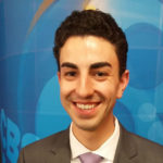 A young man with dark hair, smiling, looking at camera, with blue background.