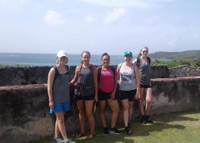 A group of females stand by stone wall, smiling, looking at camera, with cliffs and ocean in background.