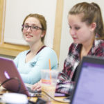 Two female students study together at a desk, using laptops.