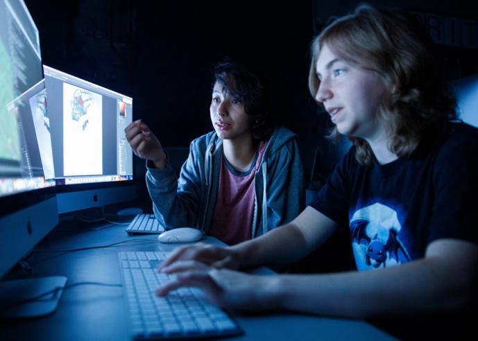 Two students work in a dark computer lab, faces lit up by computer monitors.