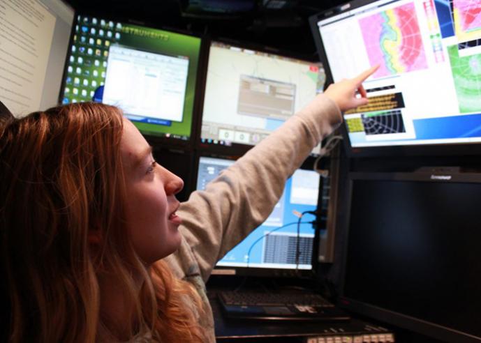 A female with red hair points to a screen showing a weather map.