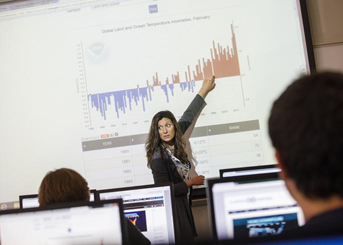 A female teacher is pointing to a graph on a projection screen to a class.