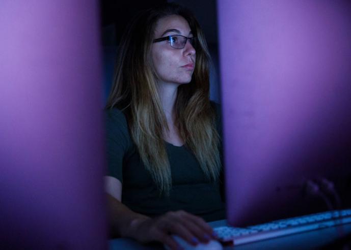 A young woman with long hair and glasses works in front of a computer screen.
