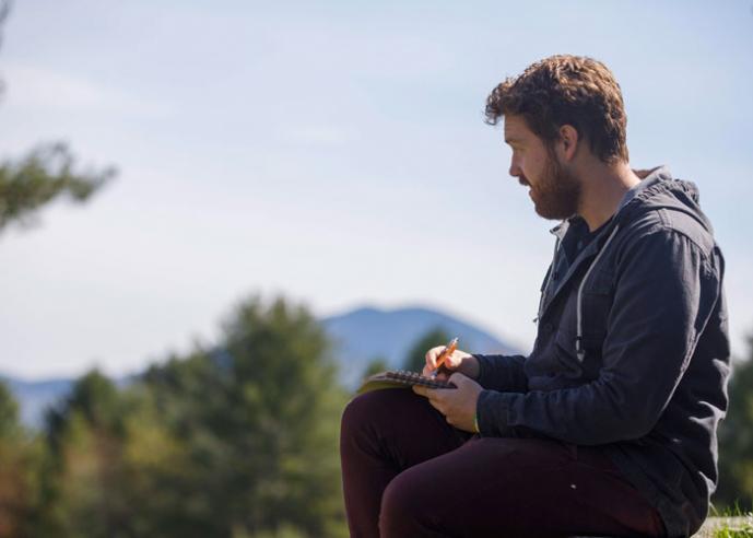 A man is drawing with a pencil on a pad, sitting outside with trees and hills in background.