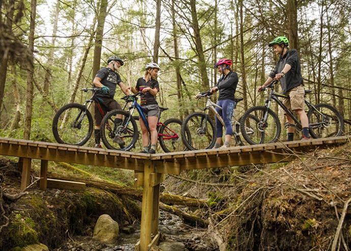 Four people wearing bike helmets, standing with their bicycles on a wooden bridge in the woods.