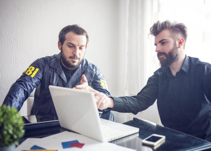 Two men with beards, one with the words FBI on his sleeve, point at a laptop while working together.