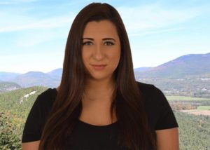 A woman with long, brown hair, looking at camera, with mountain landscape in background.