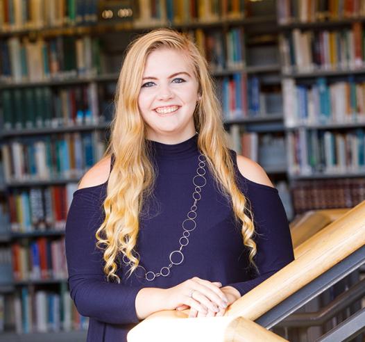 A young woman with long, blonde hair, smiling, looking at camera, with bookshelves in background.