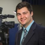 A young man with dark hair, wearing a suit, smiling, looking at camera, with video equipment in background.