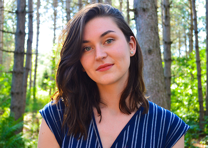 A young woman with dark hair and a blue blouse, looks into camera, with trees in background.