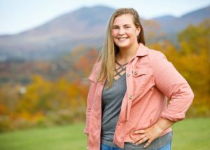 A young woman with long, blonde hair, stands with a hand on her hip, smiling at the camera, with hills and trees in background.