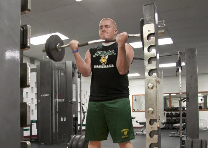 A young man wearing a black t shirt and green shorts, lifts weights in a gym.