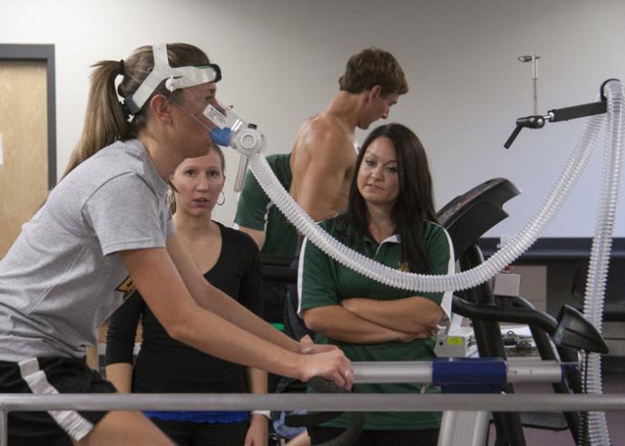 A young woman on a stationary bike in a gym wears a breathing apparatus while 2 females look on.