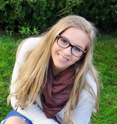 A young woman with long, blonde hair and glasses, smiling, looking at camera with grass and trees in background.