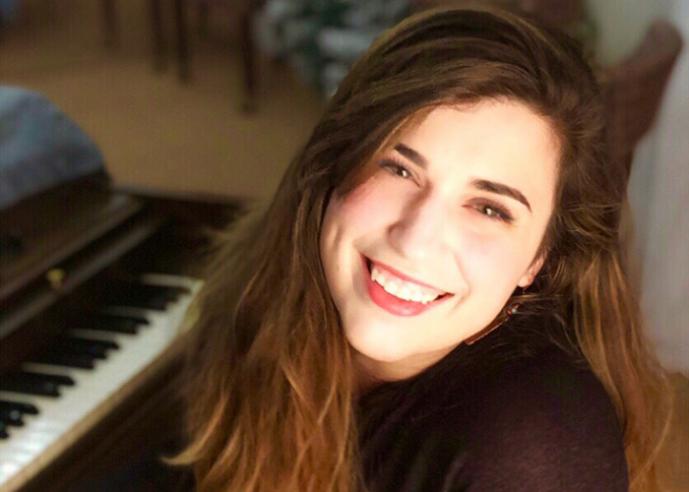 Young woman with long brown hair, smiling, looking at camera, piano in background.