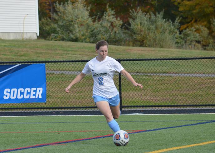 A female soccer player kicks a soccer ball on an field, fence in background.