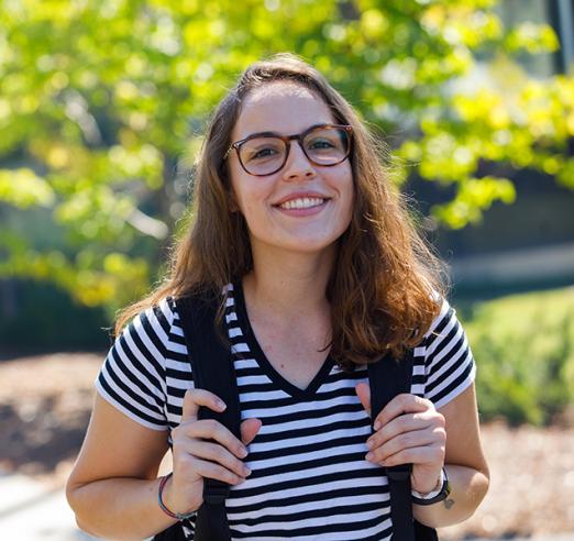 A young woman with dark hair and glasses, wearing a striped t shirt and a backpack, smiling, looking at camera.