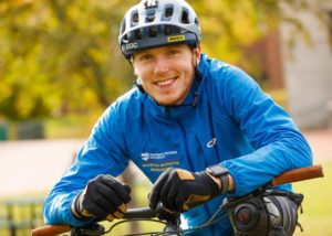 A man with a bicycle is wearing a bike helmet, a blue windbreaker and black gloves, smiling, looking at camera.