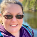 A young lady wearing sunglasses, smiling, looking at camera, with pond in background.