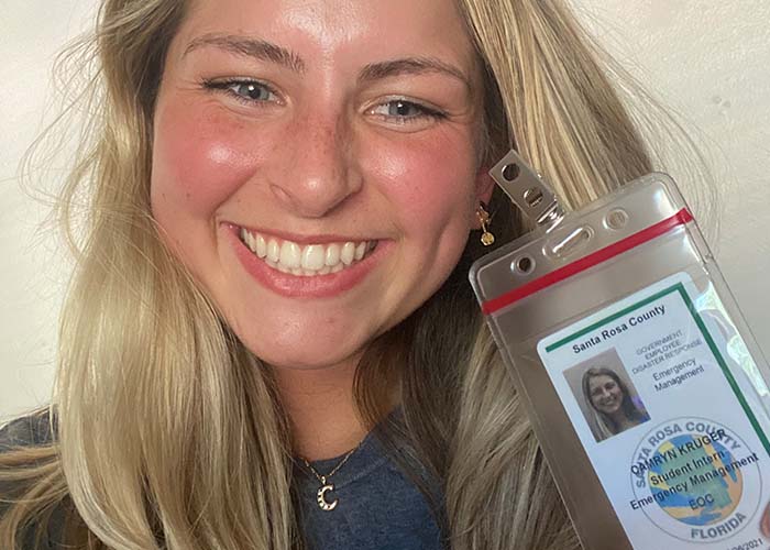 A closeup of a blond woman's face. She's smiling brightly and holding a work badge up to the camera.