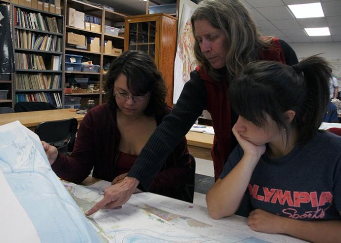 A female teacher points to a map on a desk while 2 female students look on, bookshelves in background.