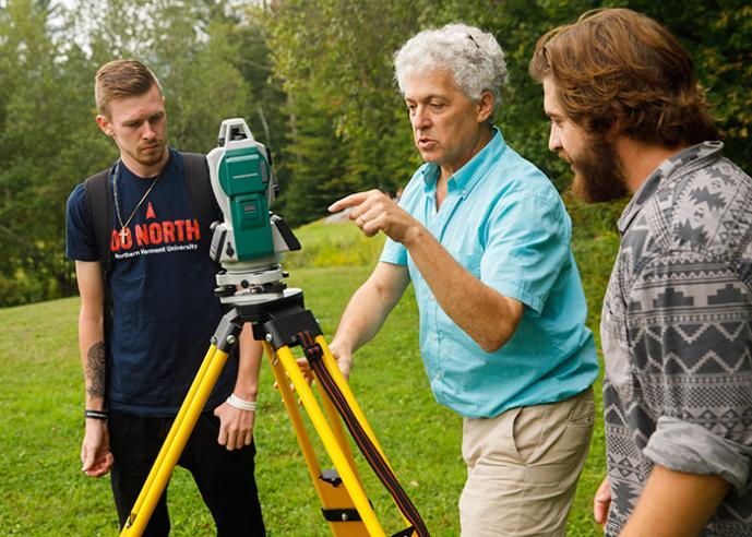 A male teacher wearing an bright blue shirt shows 2 male students survey equipment outdoors, trees in background.