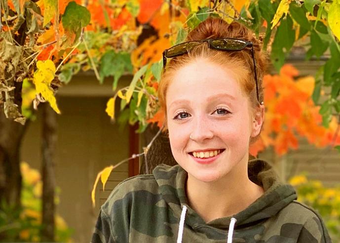 A young woman with red hair in a bun and sunglasses on her head, smiles at the camera, with autumn leaves in background.