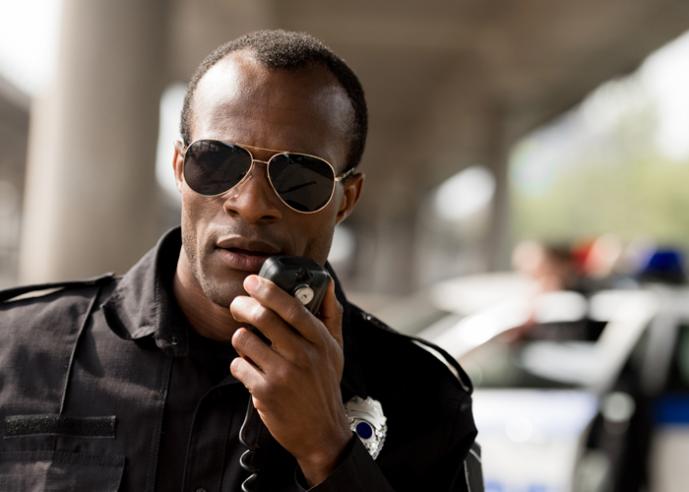 A male police officer wearing sunglasses and uniform speaks into a radio.