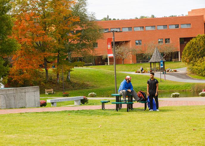 Two male students hang out at a picnic table, with trees and a campus building in background.